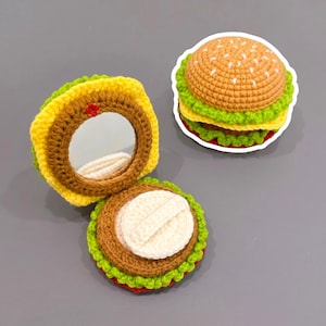 Könnte beinhalten: Ein gehäkelter Hamburger-Taschenspiegel und ein passender geschlossener gehäkelter Hamburger. Der geöffnete Spiegel zeigt eine runde Spiegelfläche, umgeben von braunen, gelben und grünen Garnschichten, mit einem weißen Puderquast im Unterteil.
