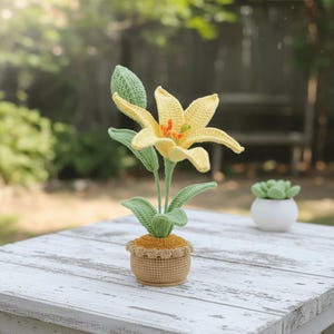 May include: A crocheted yellow lily flower with green leaves and stem, planted in a small brown pot. The flower has orange accents in the center. The pot is sitting on a white wooden table.