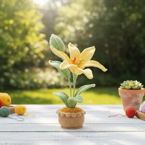 May include: A handcrafted crochet lily flower in a small pot. The flower has yellow petals, green leaves, and a green bud. The pot is tan with a scalloped edge. The background is a blurred outdoor scene with sunlight.
