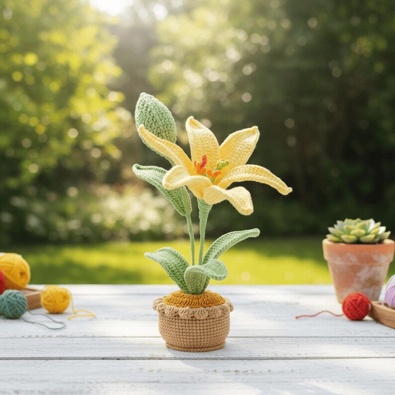May include: A handcrafted crochet lily flower in a small pot. The flower has yellow petals, green leaves, and a green bud. The pot is tan with a scalloped edge. The background is a blurred outdoor scene with sunlight.