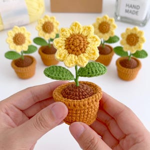 May include: A small, handmade crochet sunflower potted in a textured brown yarn pot. The flower features a dark brown centre with bright yellow petals and two green leaves on a stem. Several identical crochet sunflowers are arranged in the background.