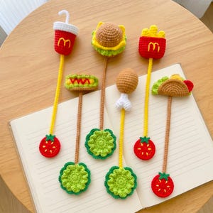 以下が含まれることがあります： 黄色いMのロゴ入りドリンクカップ、ハンバーガー、黄色いMのロゴ入りフライドポテト、チキンレッグ、タコスをモチーフにした、手編みのクロッシェ編みブックマーク5点セット。それぞれ長い茎と円形の土台が付いています。