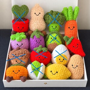 Könnte beinhalten: Eine Sammlung von gehäkelten Plüschfiguren in Form von Obst und Gemüse mit lächelnden Gesichtern, darunter eine Karotte, eine Aubergine, eine Zitrone, eine Kartoffel und weitere Produkte, angeordnet in einer weißen rechteckigen Box.