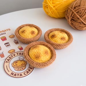 May include: Three crocheted egg tarts with golden-yellow centers and brown crusts, accompanied by a round sticker featuring a cartoon tart and the text "TASTY TREATS NATA CAFE," alongside two balls of yellow and brown yarn in the background.