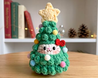 May include: A crocheted plush toy shaped like a Christmas tree with a textured green body, a yellow star topper, and small colorful pom-poms. A white cat face with black eyes, pink cheeks, and a red bow is visible in the center of the tree.