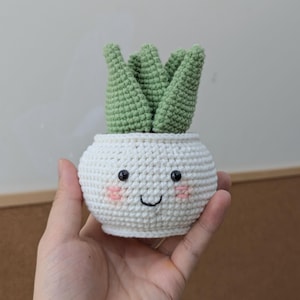 Puede incluir: Una planta suculenta de ganchillo en una maceta blanca con una cara sonriente. La planta tiene hojas verdes y la maceta tiene ojos negros, mejillas rosadas y una sonrisa cosida. Un artículo decorativo hecho a mano.