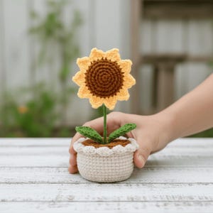 May include: A crocheted sunflower in a small, off-white pot. The sunflower has a brown center and yellow petals. Green leaves and a green stem support the flower. The pot has a scalloped edge.