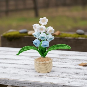 Peut inclure: Un arrangement floral crocheté avec des fleurs blanches et bleu clair en forme de cloche, des feuilles vertes et un pot beige à bord festonné. L'arrangement est posé sur une surface en bois blanc, évoquant le printemps et la nature.