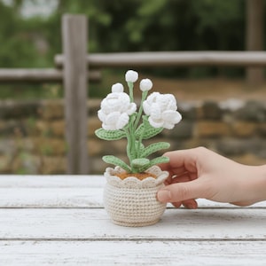 Peut inclure: Un arrangement floral crocheté avec des fleurs blanches et des feuilles vertes dans un petit pot beige. Le pot a un bord festonné. L'arrangement est posé sur une surface en bois blanc. L'arrière-plan est flou, suggérant un cadre extérieur.