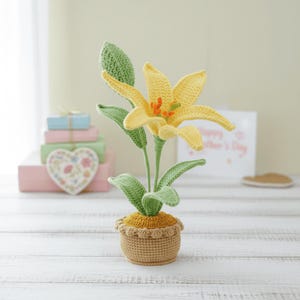 Może przedstawiać: Szydełkowy żółty kwiat lilii z zielonymi liśćmi i łodygą, w doniczce w brązowym koszu. Kwiat ma pomarańczowe akcenty pośrodku. Rzemiosło jest ustawione na białej drewnianej powierzchni.