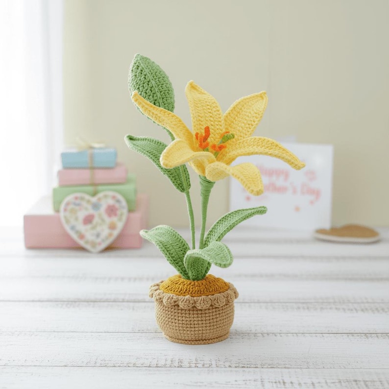 May include: A crocheted yellow lily flower with green leaves and stem, potted in a tan basket. The flower has orange accents in the center. The craft is set on a white wooden surface.