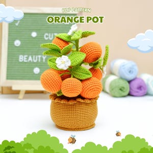 May include: A crocheted orange pot with a miniature orange tree. The tree has bright orange crocheted oranges, green leaves, and white flowers. The pot is a woven brown colour. The text "PDF PATTERN ORANGE POT" is at the top.