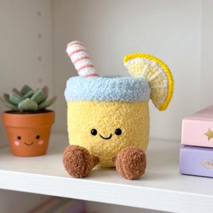 May include: A yellow crochet plush toy shaped like a drink with a light blue rim, a striped straw, and a yellow lemon slice garnish. The plush has two black eyes, a stitched smile, and two brown feet. Beside it sits a small terracotta-coloured planter.