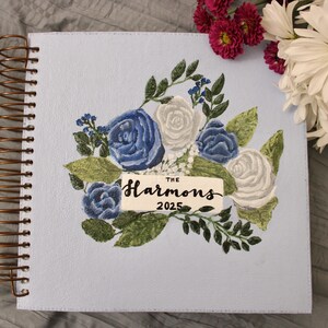 Puede incluir: Un cuaderno azul claro con espiral y diseño floral. La portada presenta rosas azules y blancas, hojas verdes y el texto "The Harmons 2025". La encuadernación en espiral es de color cobre.