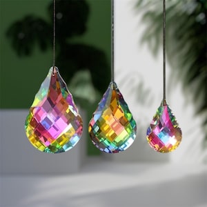 May include: Three teardrop-shaped crystal prisms hanging from thin black strings. Each faceted glass piece displays a vibrant rainbow spectrum of colours, including pink, yellow, green, and blue, reflecting light across their geometric surfaces.
