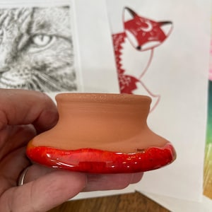 May include: A small, circular terracotta pottery piece with a flared base and a smooth, unglazed tan upper section. The wide, rounded bottom rim is finished with a vibrant, glossy red glaze that contrasts with the natural clay texture above.