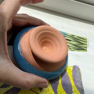 May include: A handmade ceramic bowl with a terracotta interior and a speckled blue exterior. The bowl has a unique, layered design. The bowl is held by a hand.