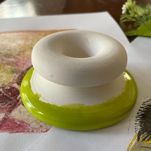 May include: A handmade ceramic object with a white, donut-shaped top and a bright green base. The base is a glossy, circular shape. The object is sitting on a white surface with colorful artwork in the background.
