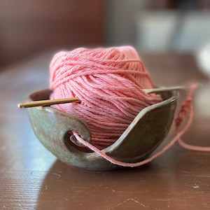 May include: A ceramic yarn bowl in a muted green and brown glaze holds a large ball of pink yarn. A wooden crochet hook rests across the top. The yarn's end feeds through a swirl-shaped opening in the bowl's side.
