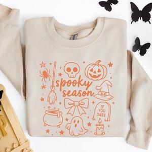 May include: A beige sweatshirt with an orange graphic that says "spooky season" and features Halloween-themed illustrations. The design includes a spider, skull, pumpkin, witch's hat, and other festive elements. Black butterfly decorations and a skull are in the background.