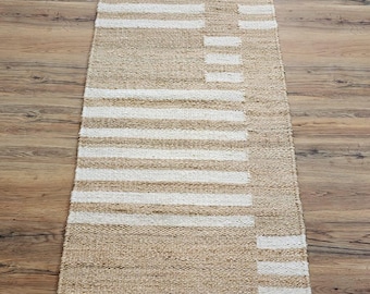 May include: A rectangular jute rug with a geometric pattern. It has alternating horizontal stripes of natural tan and white. A vertical white stripe intersects the horizontal stripes, creating a unique design. The rug is on a wooden floor.