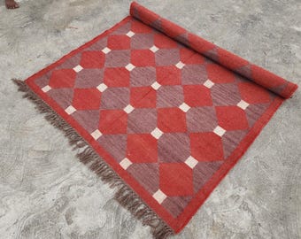May include: A red and grey checkered rug with a rolled-up section. The rug features a geometric pattern of alternating red and grey squares with white squares in the centre of each grey square. The rug has a fringe on one edge.