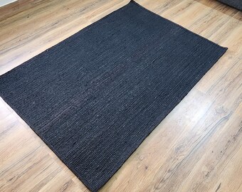 May include: A rectangular, dark gray woven rug. The rug is placed on a light brown wooden floor. The rug's texture is visible, with a slightly uneven surface. The rug is a solid color and appears to be made of natural fibers.