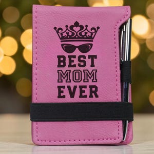 May include: Pink notepad with the text "BEST MOM EVER" in black, featuring a crown and sunglasses graphic. A black elastic band secures the notepad, and a pen is inserted in a side loop. The background is blurred with bokeh lights.