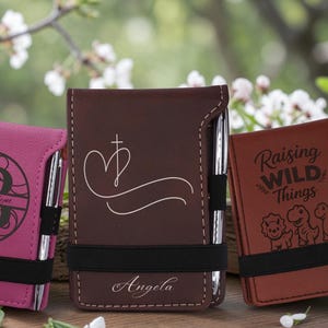 May include: Three rectangular pocket notebooks with elastic pen loops and silver pens. The pink notebook features a monogram with the name Brooklyn. The dark brown notebook displays a heart cross design and the name Angela. The tan notebook says Raising Wild Things with dinosaur illustrations.