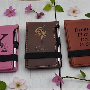 May include: Three personalized notepad holders in pink, brown, and tan leather. Each notepad holder has a pen, a black elastic band, and unique designs: a floral initial, a floral design with a name, and the text "Dream. Plan. Do."