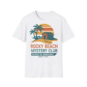 Könnte beinhalten: Ein weißes T-Shirt mit kurzen Ärmeln und einer Grafik, die einen Strand mit Palmen, einer kleinen Hütte und einem Sonnenuntergang zeigt. Der Text lautet ROCKY BEACH MYSTERY CLUB, SOLVING THE UNSOLVABLE und SINCE 1964 in Orange und Petrol.