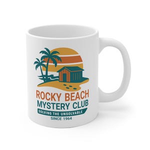 Op de afbeelding: Witte keramische mok met een strandscène: palmbomen, een klein houten gebouw en een zonsondergang. De tekst "Rocky Beach Mystery Club" en "Solving the Unsolvable Since 1964" staat op de mok.