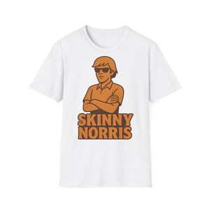 Könnte beinhalten: Ein weißes kurzärmeliges T-Shirt mit einer grafischen Illustration einer Person mit braunen Haaren, die eine Sonnenbrille und ein Hemd mit Kragen trägt und die Arme verschränkt hat. Unter der Figur steht der Text SKINNY NORRIS in braunen Buchstaben.