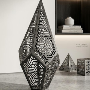 May include: A tall, dark gray, diamond-shaped sculpture featuring an intricate, laser-cut geometric lattice pattern. The structure is composed of multiple connected panels secured with visible rivets, creating a complex, repeating angular design.