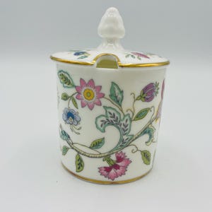 May include: A white ceramic sugar bowl with a lid and a floral design. The bowl features pink, blue, and purple flowers with green leaves. The rim and base are trimmed with gold. The lid has a small handle.