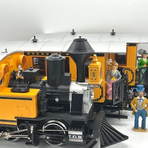 May include: A detailed model train set featuring a yellow and black steam engine with the text "LGB" and a passenger car. Miniature figurines of people are positioned around the train, adding a sense of scale and detail.