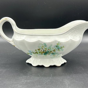 May include: A white porcelain gravy boat with a handle and spout. The boat features a floral design in shades of blue and green, with gold trim accents. The base has a scalloped edge.