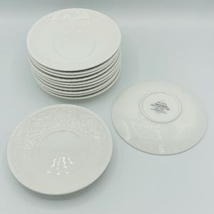 May include: A stack of white saucers with a floral embossed design. Two additional saucers are displayed, one face up and one face down, showcasing the embossed pattern and the brand name "Mikasa Maxima". The saucers are made of a glossy white material.