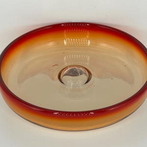 Puede incluir: Un cuenco de vidrio transparente con forma circular y un pequeño centro elevado. El borde del cuenco tiene un degradado de colores, que va del rojo intenso al naranja claro. El cuenco probablemente se utiliza con fines decorativos.
