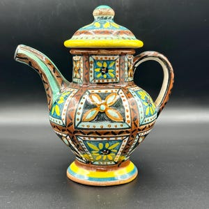 May include: A decorative ceramic teapot with a lid. The teapot features a colorful geometric pattern in shades of brown, yellow, blue, and green. The spout and handle are curved. The teapot sits on a small, round base.