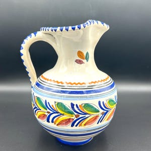 May include: A white ceramic pitcher with a curved handle and spout. The pitcher is decorated with blue, orange, yellow, and green floral patterns and stripes. The rim and base are blue, and the handle has a textured design.
