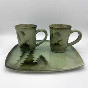 May include: Two green ceramic mugs with handles and a matching tray. The mugs and tray feature a forest and moon design. The set is a muted green color with darker green accents.