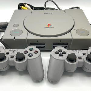 May include: A gray Sony PlayStation console with two matching controllers. The console has a disc tray, power and open buttons, and the PlayStation logo. The controllers have analog sticks and directional buttons. Cables are connected to the console.