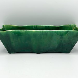 May include: A rectangular, emerald green ceramic planter with a glossy finish. The planter has a unique, angular design with slightly flared sides and a wavy top edge. The planter is empty and sits on a white surface.