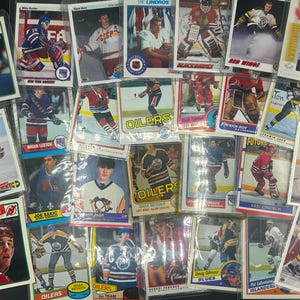 May include: A collection of vintage hockey trading cards featuring various players and teams. The cards are in protective sleeves and showcase action shots and portraits. Teams include Oilers, Red Wings, and Islanders.
