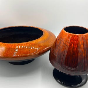 May include: Two ceramic vases with a glossy finish. One is a wide, shallow bowl in shades of orange and brown. The other is a rounded vase in deep red and brown tones, set on a black base. Both have black interiors.