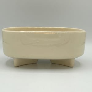 May include: Cream-colored ceramic planter with a rectangular shape and three small, square feet. The planter has a smooth, glossy finish and is suitable for small plants or succulents. The neutral color makes it versatile for various decor styles.