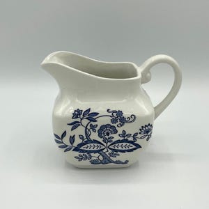 May include: A white ceramic creamer with a blue floral pattern. The creamer has a square base, a curved handle, and a spout for pouring. The blue floral design includes flowers, leaves, and vines. The creamer measures approximately 4 inches in height.