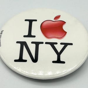 May include: Oval-shaped button with the text "I NY" in black, with a red apple logo replacing the word "love". The button has a white background and a glossy finish. The text "TM/© 2001 NYSDED" is printed on the left side.