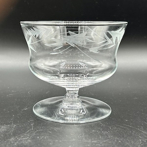 May include: A clear glass dessert dish with a wide bowl and a short stem. The bowl has an etched floral design around the rim. The dish sits on a round base. The glass is transparent, allowing light to pass through.